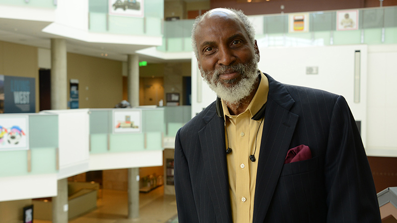 Portrait of an older man with a white beard, wearing a navy pinstriped blazer and yellow shirt, standing in a bright, multi-level building lobby.