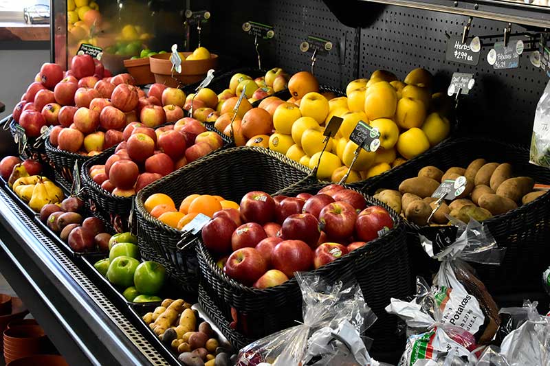 A well-stocked produce display at a small grocery store, featuring fresh apples, pears, oranges, potatoes, and other fruits and vegetables in black wicker baskets.