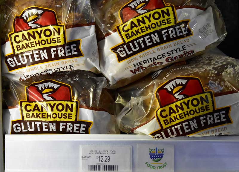 Canyon Bakehouse Gluten Free Heritage Style 100% Whole Grain Bread loaves on a store shelf, priced at $12.29, with a Food Trust price tag label.