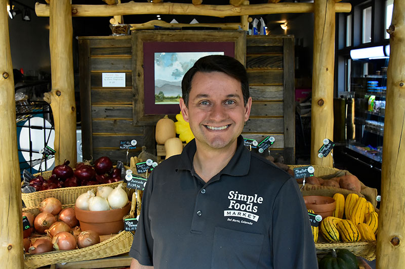 A smiling Simple Foods Market employee stands in front of a rustic wooden produce display at the Del Norte, Colorado store.