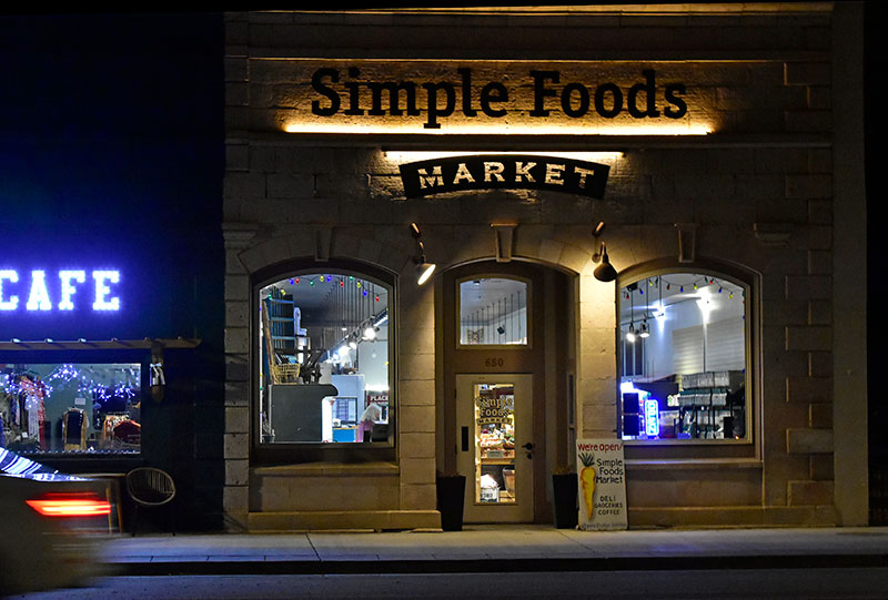 La fachada de piedra de Simple Foods Market por la noche, con luces cálidas iluminando el letrero del frente de la tienda y ventanas arqueadas brillando por dentro.