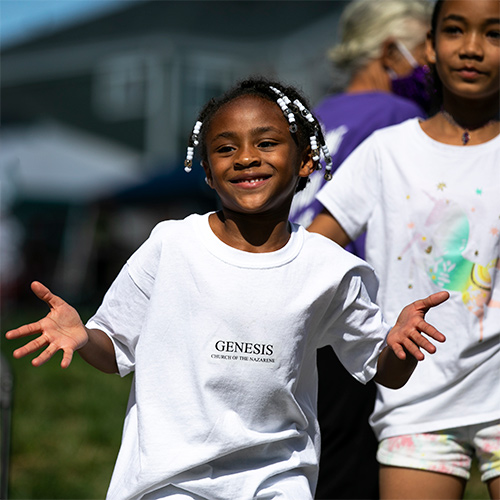 A young girl with beaded braids smiles and reaches out with both hands at an outdoor community event, wearing a white Genesis Church of the Nazarene T-shirt.